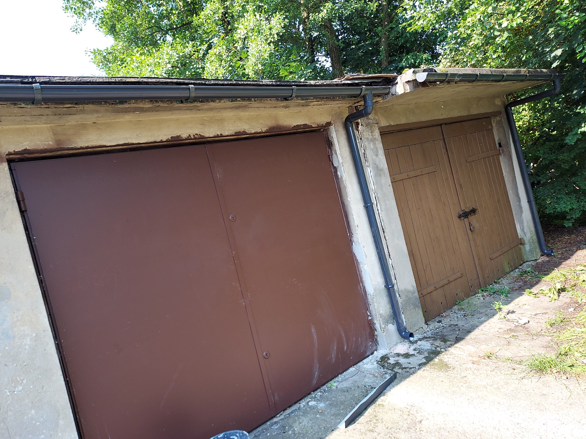 Dwa garaże z betonową konstrukcją i zamontowanymi, ciemnymi rynnami; jeden z brązową, metalową bramą, drugi z drewnianą bramą dwuskrzydłową.