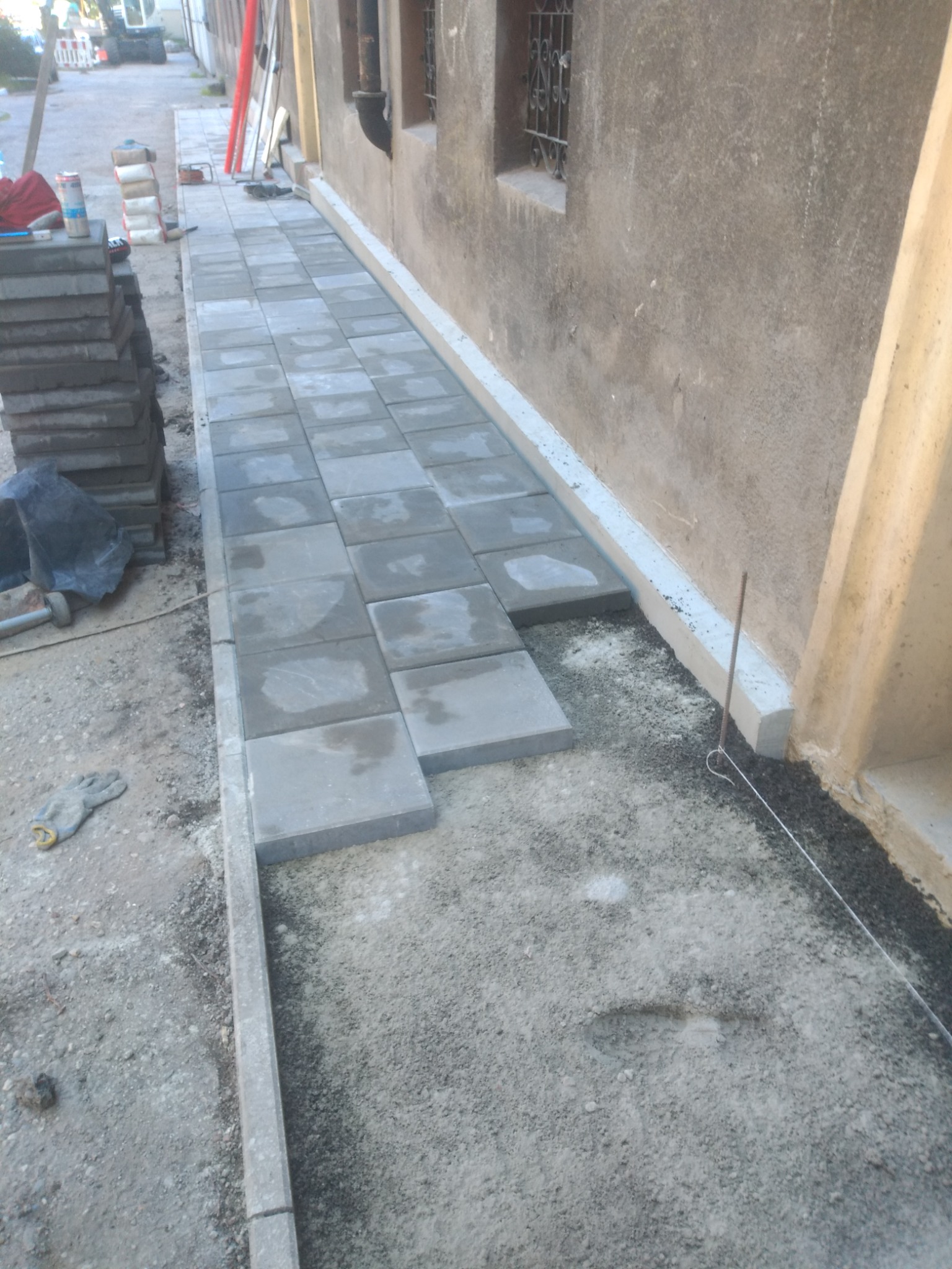 Układanie szarej kostki brukowej wzdłuż ściany budynku, widoczne narzędzia i materiały budowlane, częściowo ułożona nawierzchnia.