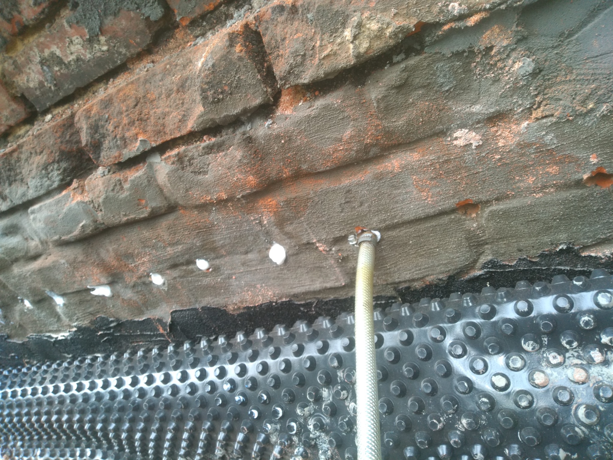 Szczegółowe ujęcie fragmentu murowanej ściany z widocznymi otworami iniekcyjnymi, pokrytej szarą zaprawą, z zamontowaną folią kubełkową i wystającą rurką iniekcyjną.