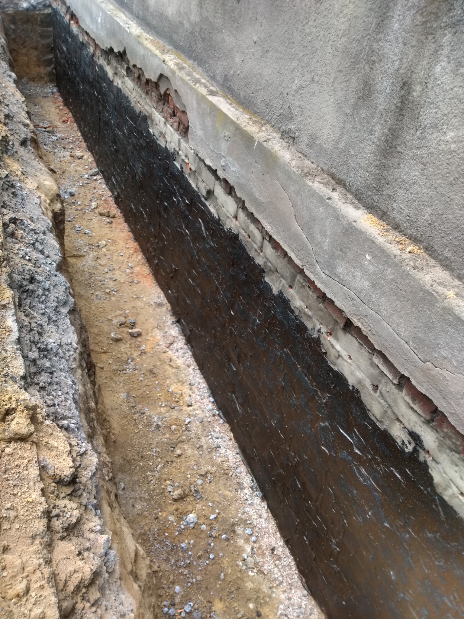 Odkopana ściana fundamentowa z cegły, zabezpieczona czarną izolacją bitumiczną, widoczny wykop w ziemi.