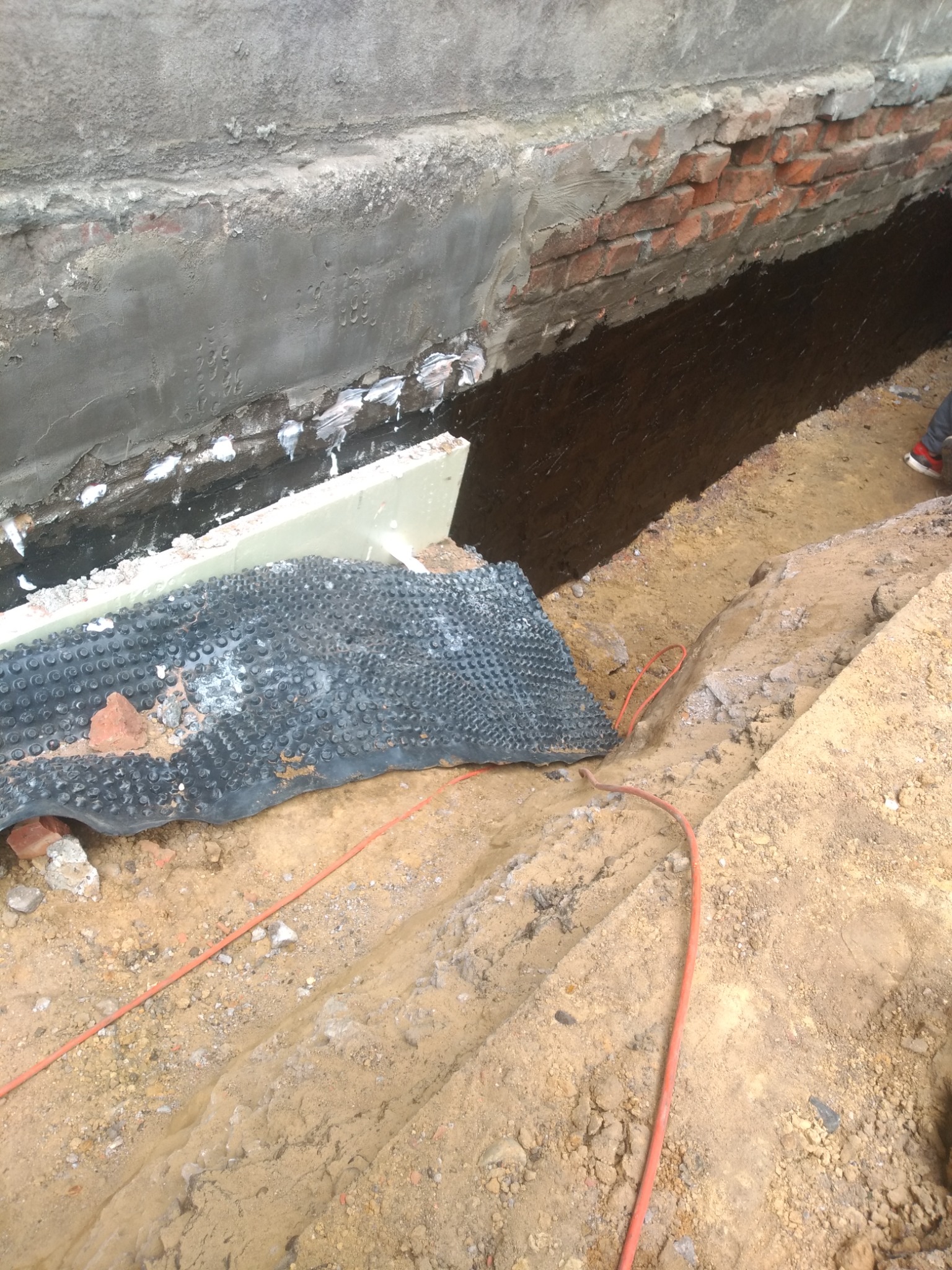 Fundament budynku w trakcie izolacji: czarna folia kubełkowa i biały styropian przy ścianie z cegły i betonu, widoczny wykop w ziemi i czerwony kabel.