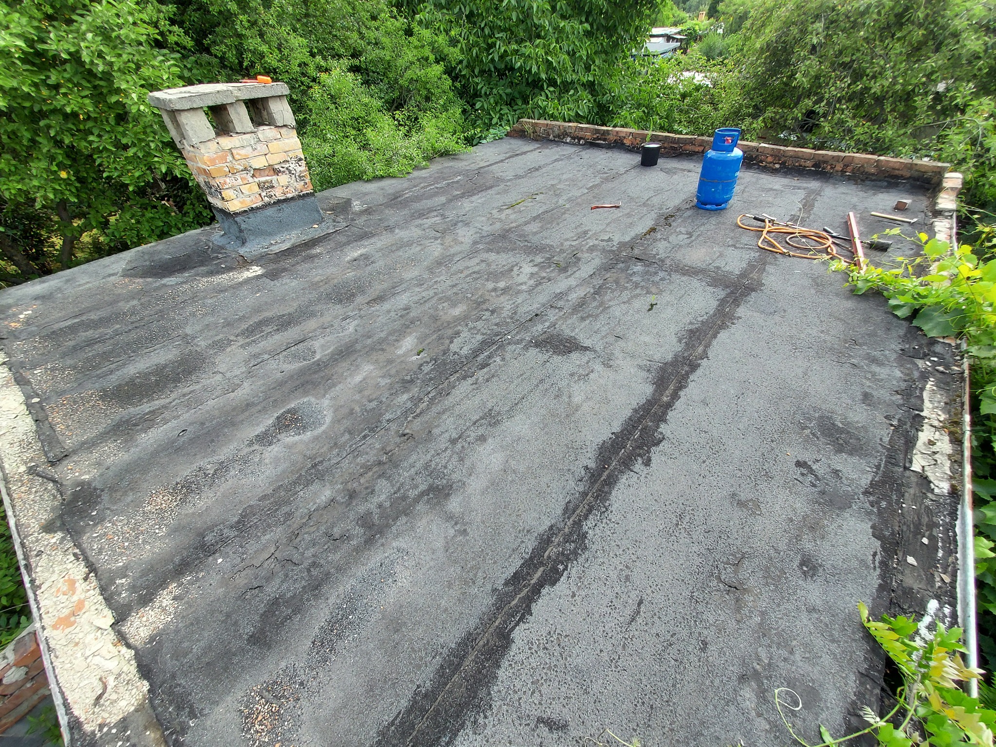 Widok z góry na płaski dach pokryty papą z ceglanym kominem, butlą gazową i narzędziami dekarskimi w otoczeniu zieleni.