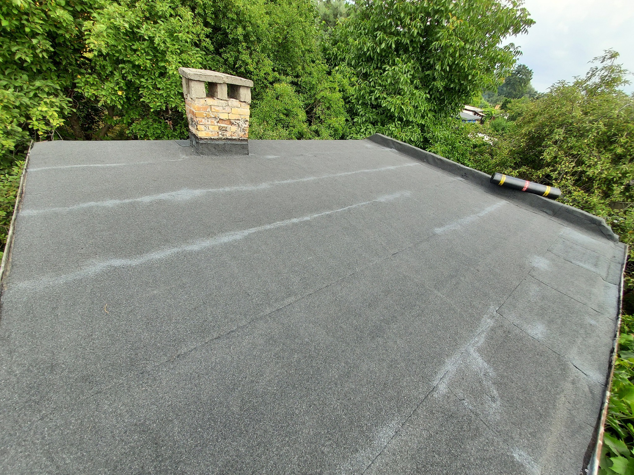 Nowy, czarny dach pokryty papą termozgrzewalną, z widocznym kominem z cegły i zielonym otoczeniem drzew.