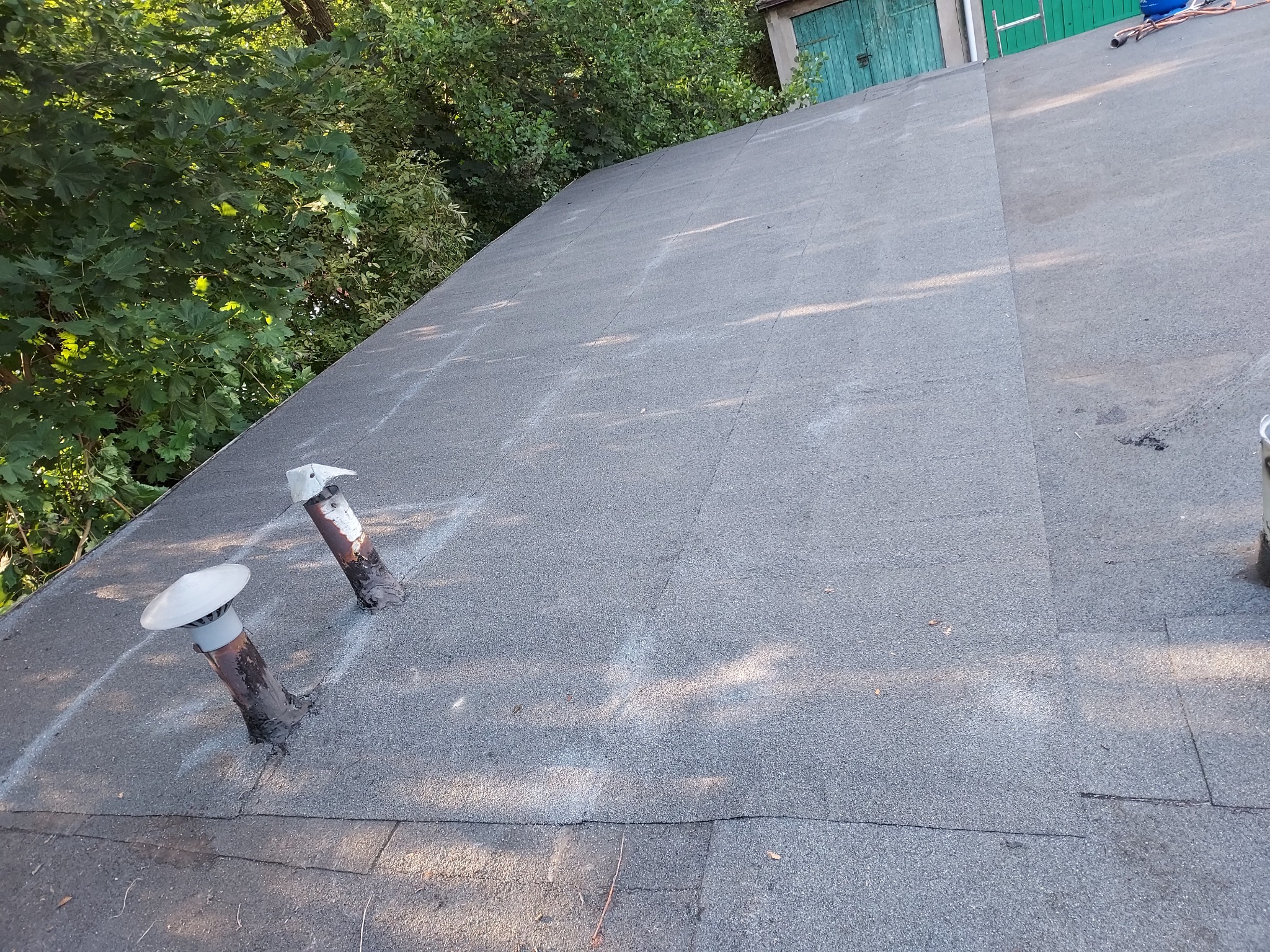 Bitumiczny dach z dwoma kominkami wentylacyjnymi na tle zieleni, widoczne ślady po łatach.