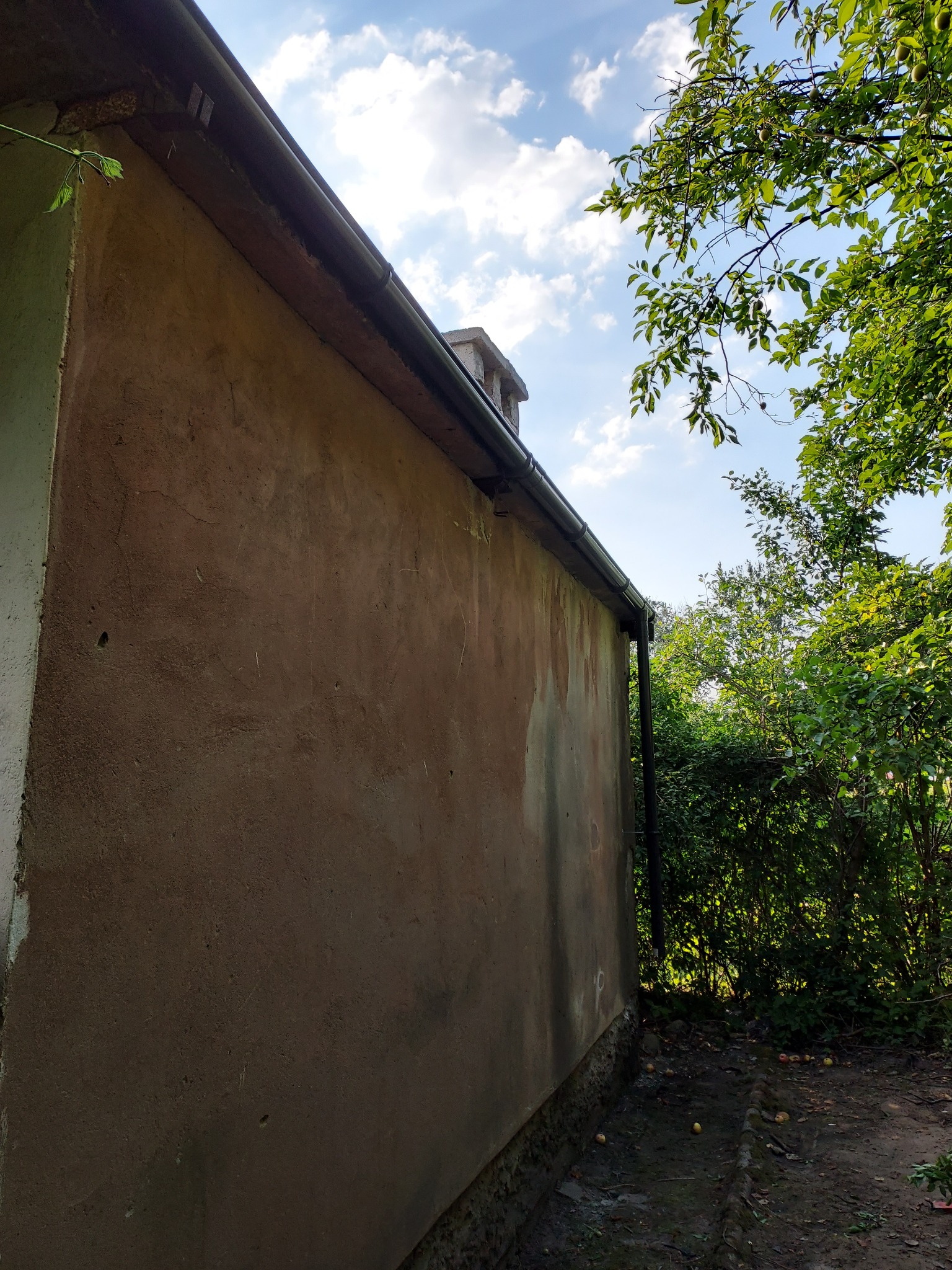 Boczny widok ściany budynku z zamontowaną rynną, widoczne pęknięcia i ubytki w elewacji, zieleń drzew w tle.