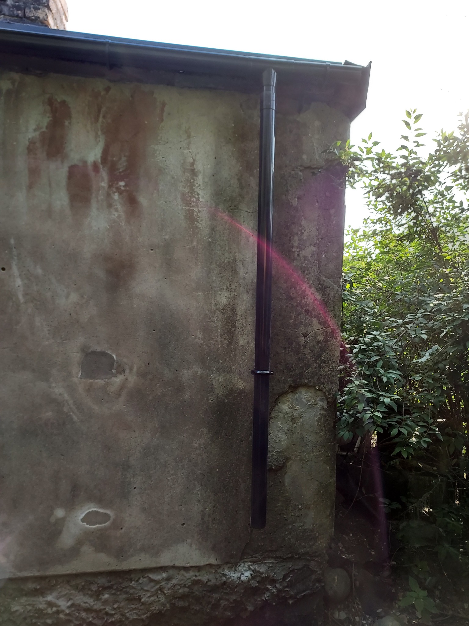 Szara, zniszczona ściana budynku z zamontowaną czarną rynną spustową, widoczne fragmenty zieleni w tle.