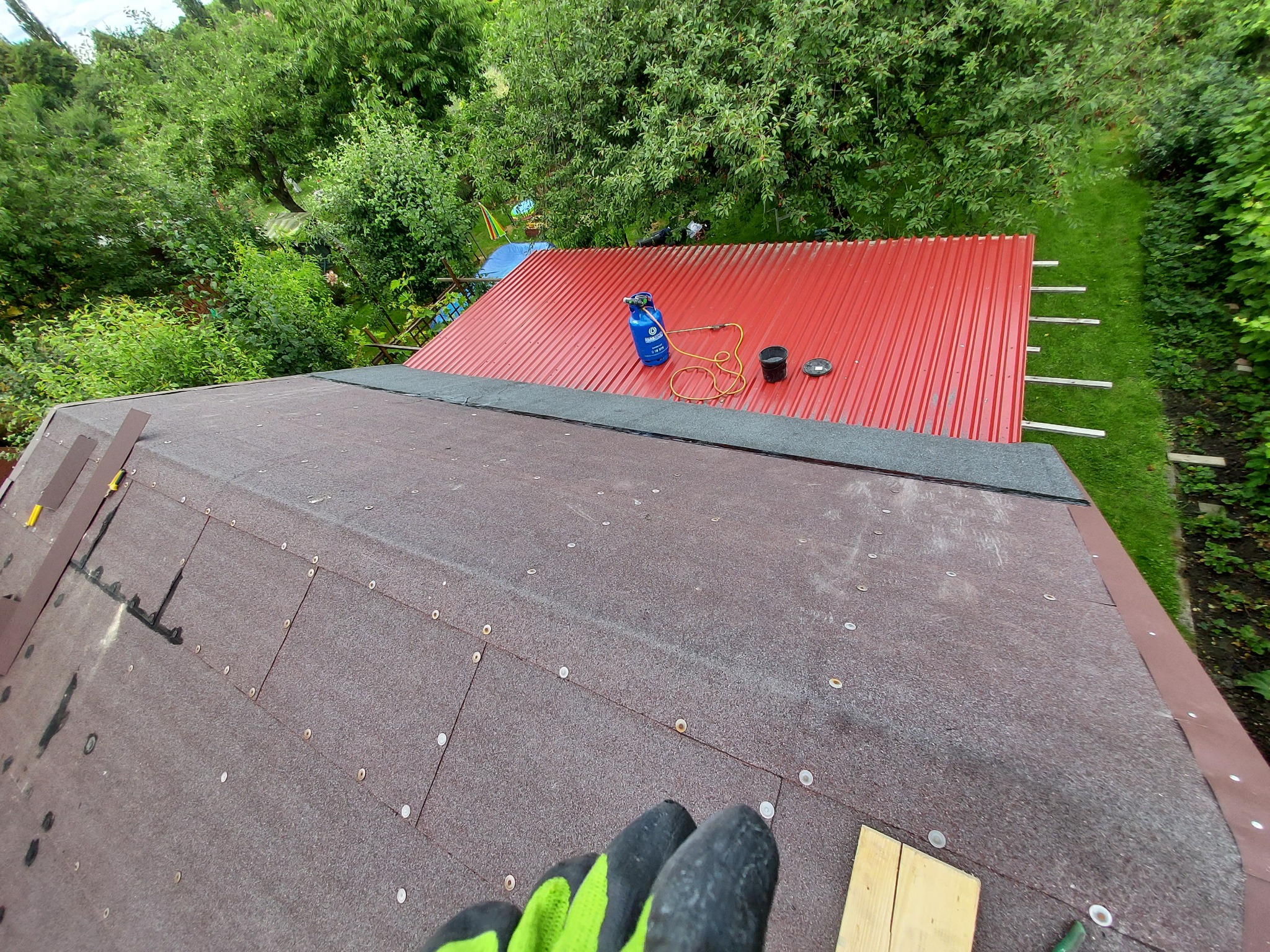 Widok z góry na fragment dachu z papy z widocznymi mocowaniami, obok blaszany dach pomalowany na czerwono z butlą gazową i narzędziami dekarskimi, w tle zieleń drzew.