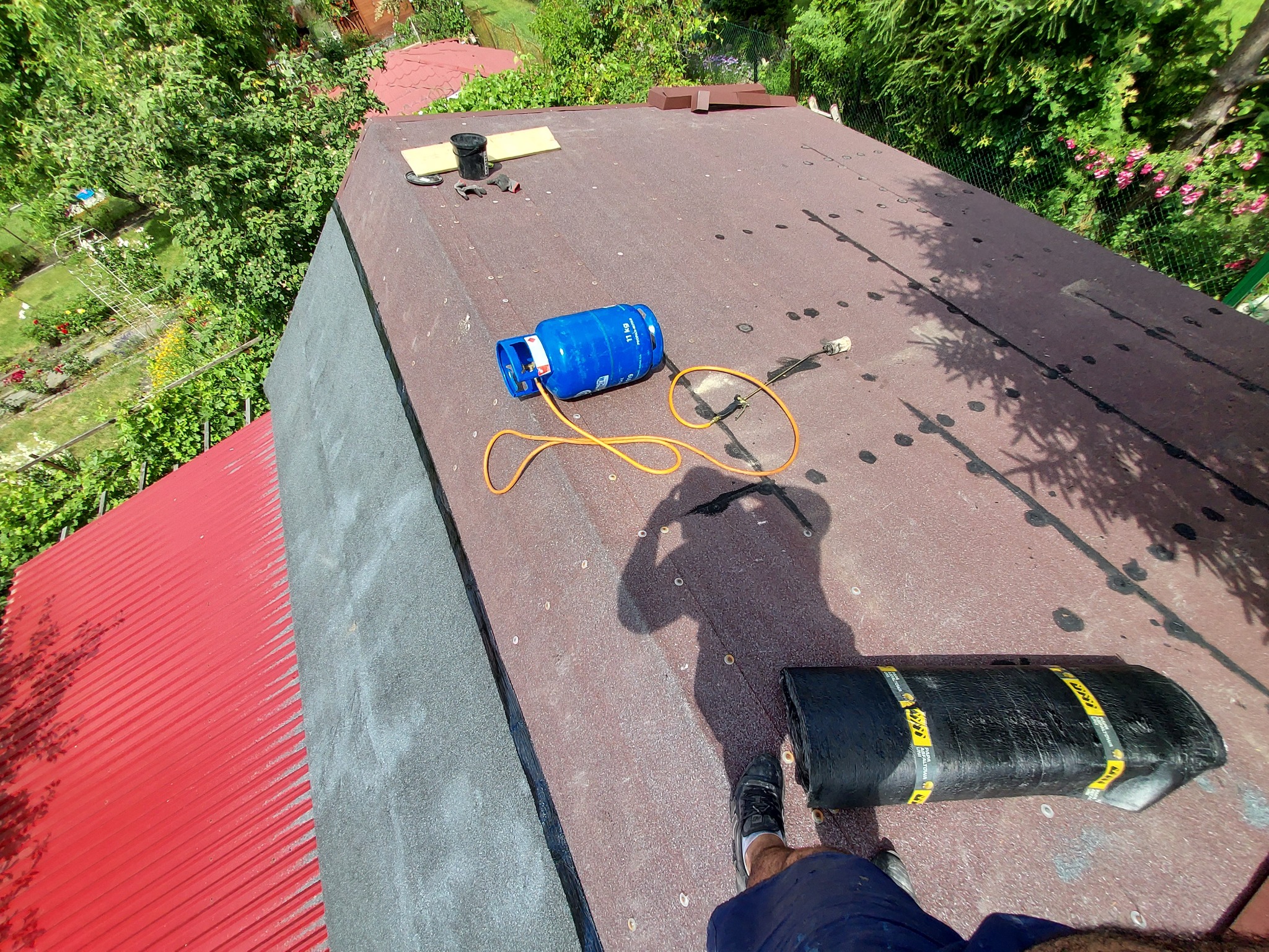 Niebieska butla gazowa i zwój papy na brązowym dachu z bitumicznym pokryciem, cień osoby pracującej, w tle zieleń drzew i czerwony dach sąsiedniego budynku.