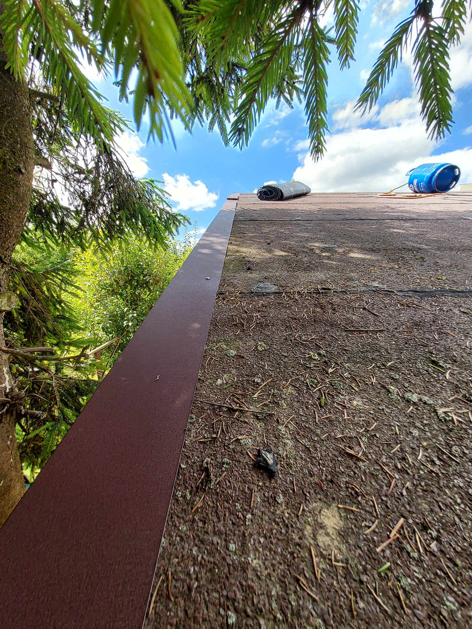 Widok z dołu na krawędź dachu z brązowym wykończeniem, z widocznymi igliwiem, drzewem i błękitnym niebem z chmurami w tle, oraz zwiniętą papą i niebieską butlą gazową na dachu.