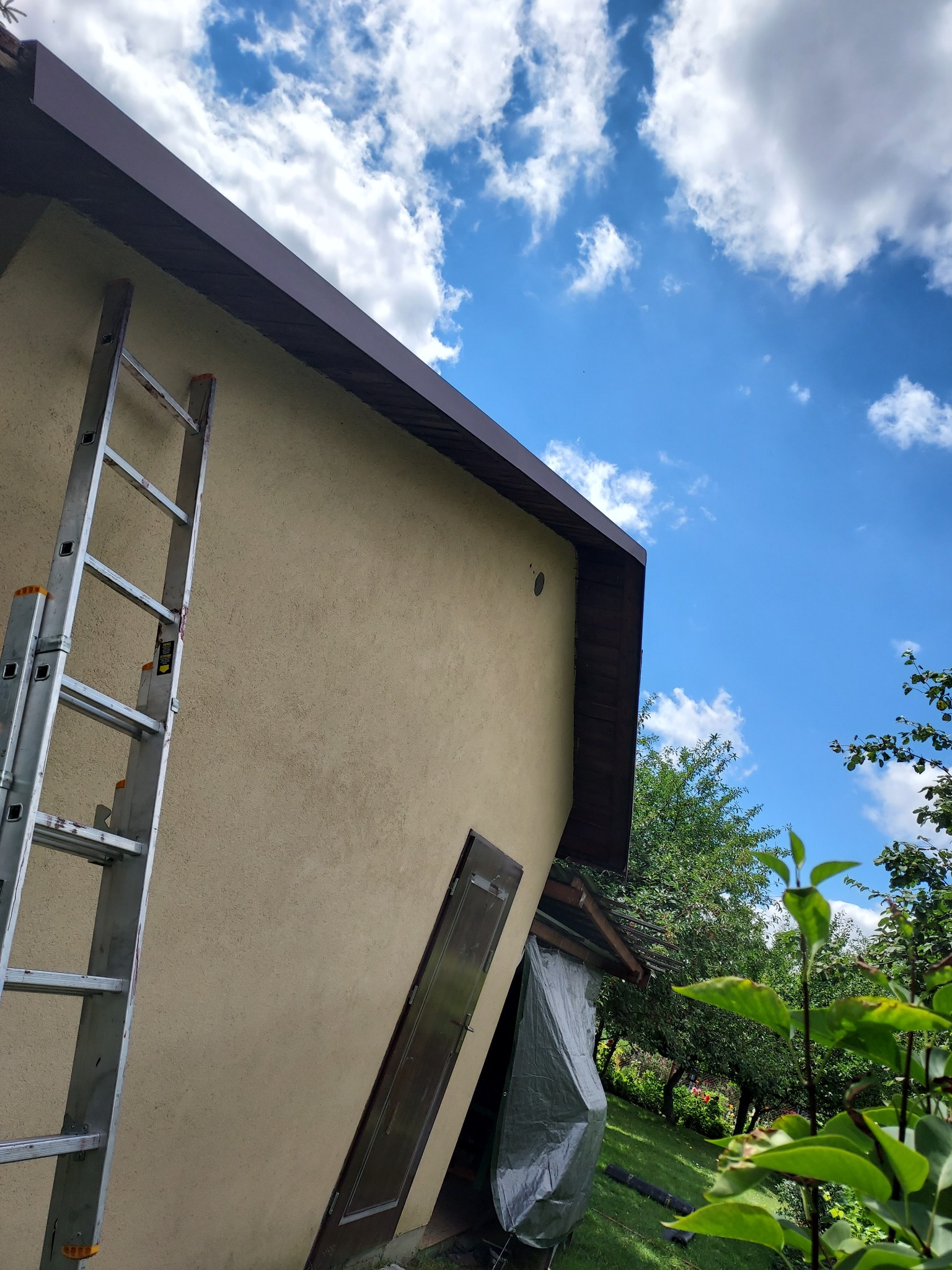 Ściana budynku z beżowym tynkiem, brązowe obramowanie dachu, metalowa drabina oparta o ścianę, błękitne niebo z białymi chmurami w tle, drzwi na ścianie.