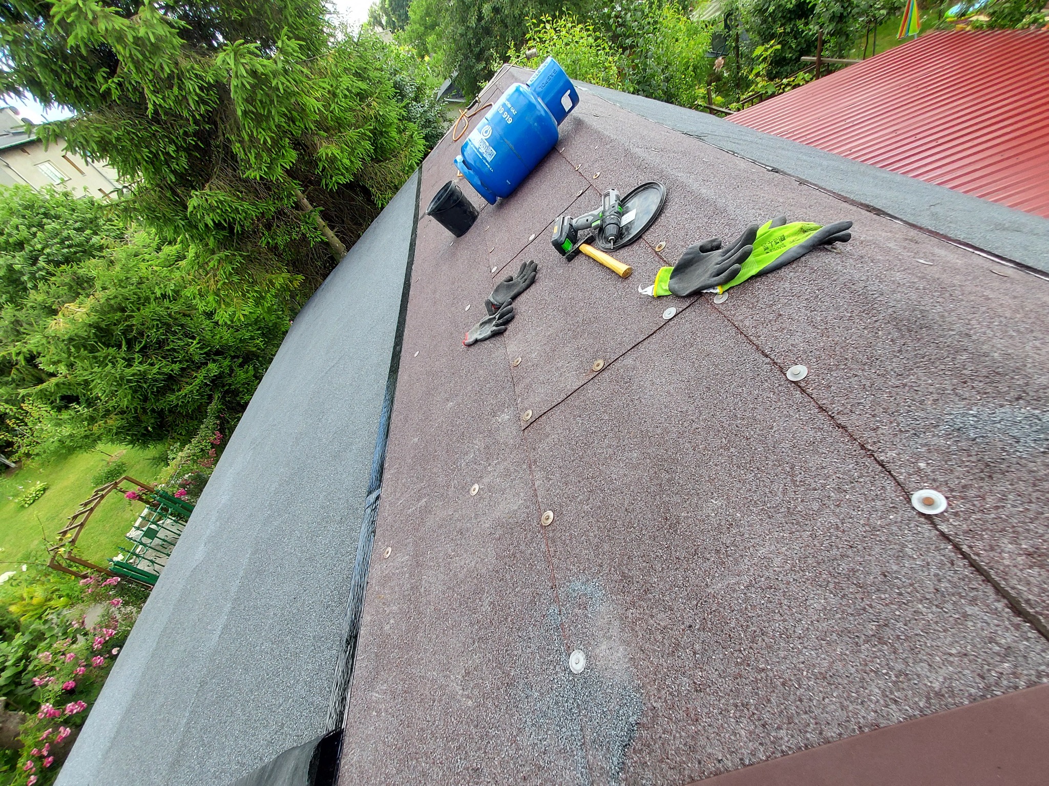 Fragment dachu pokrytego papą z widocznymi elementami mocującymi, narzędzia pracy, rękawice robocze oraz butla gazowa na tle zieleni ogrodowej.