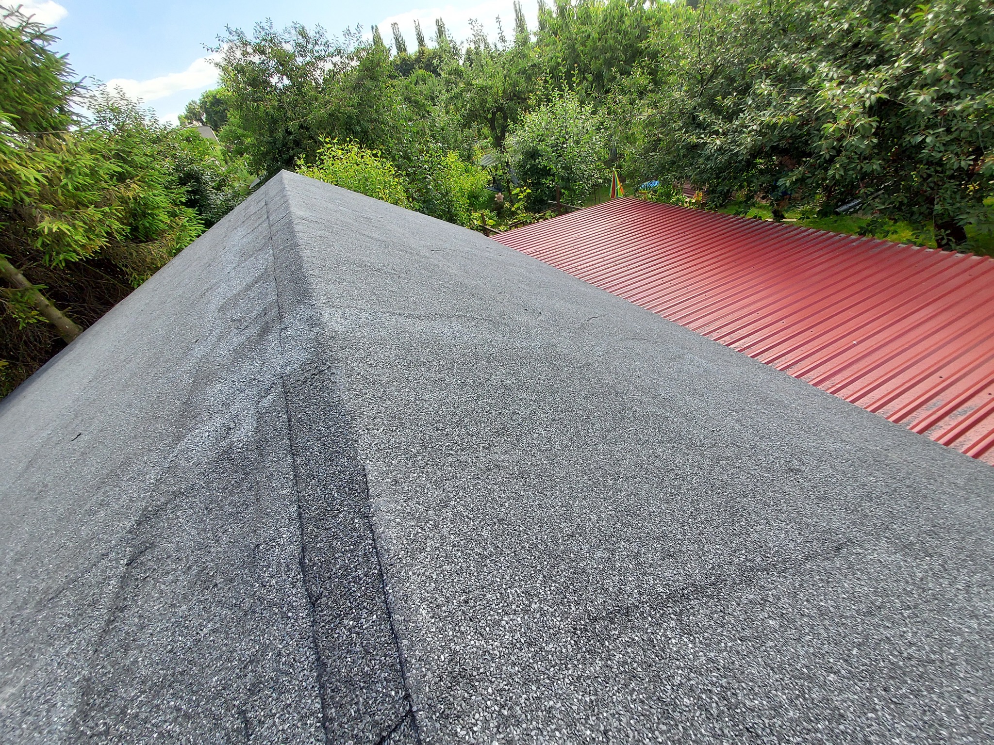 Widok z góry na fragment dachu pokrytego papą bitumiczną w kolorze grafitowym, sąsiadujący z dachem z blachy trapezowej w kolorze czerwonym, na tle zieleni drzew