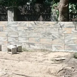 Wznoszenie murku oporowego z betonowych bloczków o marmurkowym wzorze, widoczne niedokończone elementy konstrukcji i teren budowy.