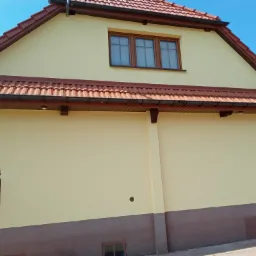 Odnowiona elewacja budynku z nową, jasną farbą, widoczne brązowe okna i dachówka, dolna część elewacji w kolorze brązowym, fragment ogrodzenia z metalowymi zdobieniami po lewej.