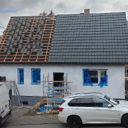 Dom jednorodzinny w trakcie prac dekarskich: połowa dachu pokryta czarną dachówką, druga połowa z widoczną konstrukcją więźby i częściowo ułożoną dachówką. Okna zabezpieczone folią, na podjeździe...