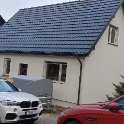 Nowy, grafitowy dach z dachówki ceramicznej na jasnym domu jednorodzinnym z widocznymi dwoma samochodami osobowymi na podjeździe.