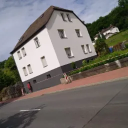 Dwupiętrowy dom z białymi ścianami i szarym cokołem, częściowo odnowiona elewacja, zabezpieczony taśmą ostrzegawczą, widok z poziomu ulicy.