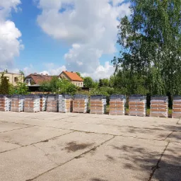 Rząd palet z workami opału na betonowym placu, w tle budynki mieszkalne i drzewa pod błękitnym niebem z chmurami.