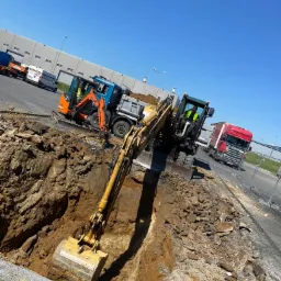 Żółta koparka w trakcie wykonywania wykopu w ziemi, obok stoi pomarańczowa mini-koparka na lawecie, a w tle widoczny budynek przemysłowy i czerwona ciężarówka.