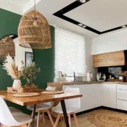 Jasne wnętrze jadalni z drewnianym stołem na czarnych nogach, białymi krzesłami, jutowym dywanem i plecioną lampą wiszącą, ściana pomalowana na ciemną zieleń.