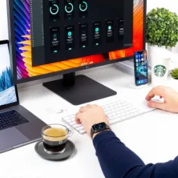 Biurko z komputerem wyświetlającym interfejs aplikacji mobilnej, obok laptop, telefon, kawa i ręka na klawiaturze.