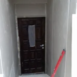 Klatka schodowa w trakcie remontu, ściany obłożone płytami gipsowo-kartonowymi, widoczny poziomica, narzędzia i materiały budowlane na górnej półce nad drzwiami.