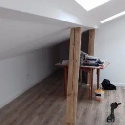 Poddasze po remoncie: białe ściany, drewniane belki konstrukcyjne, brązowe panele podłogowe, okno dachowe, drabina i narzędzia malarskie na podłodze.