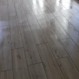 Połyskująca, jasna podłoga z płytek imitujących drewno ułożonych w regularny wzór, widok z niskiej perspektywy w kierunku okna balkonowego, ściany w tle przygotowane do malowania.