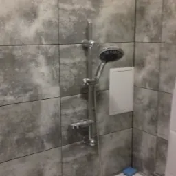 Szare płytki imitujące beton w kabinie prysznicowej z białą wanną i niebieską gąbką w rogu.