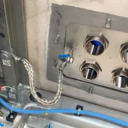 Metalowa skrzynka elektryczna z licznymi otworami i podłączonymi przewodami uziemiającymi, widoczne niebieskie i czerwone kable oraz oznaczenia uziemienia.