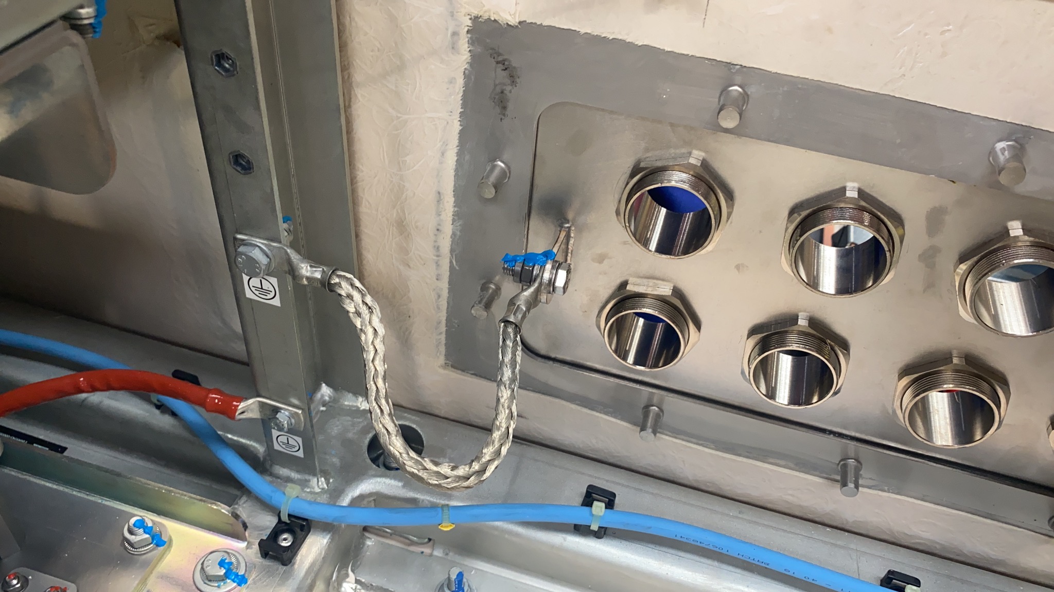 Metalowa skrzynka elektryczna z licznymi otworami i podłączonymi przewodami uziemiającymi, widoczne niebieskie i czerwone kable oraz oznaczenia uziemienia.