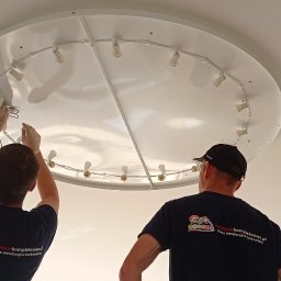 Instacjekompleksowo.pl- Tze.pl Wrocław - Dwóch elektryków w koszulkach firmowych instaluje okrągłą lampę sufitową z wieloma żarówkami, jeden z nich w rękawicach łączy przewody.