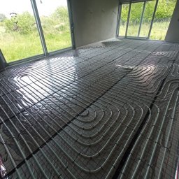 Instacjekompleksowo.pl- Tze.pl Wrocław - Białe rury ogrzewania podłogowego ułożone na folii izolacyjnej z czarnymi liniami siatki w surowym wnętrzu z dużymi oknami i widokiem na zieleń.