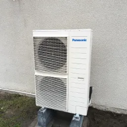 Zewnętrzna jednostka klimatyzacji Panasonic, zamontowana na betonowych podstawach przy szarej ścianie budynku z widoczną rurą spustową i kratką wentylacyjną.