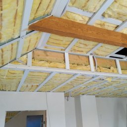 Instacjekompleksowo.pl- Tze.pl Wrocław - Sufit podwieszany z izolacją z wełny mineralnej, widoczne profile metalowe, drewniana belka i drabina oparta o ścianę w trakcie montażu.