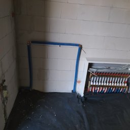 Instacjekompleksowo.pl- Tze.pl Wrocław - Rozdzielacz ogrzewania podłogowego wnękowy, rury niebieskie prowadzone wzdłuż ściany z bloczków, czarna folia izolacyjna na podłodze, surowy stan wykończenia.