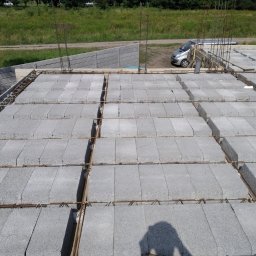 Instacjekompleksowo.pl- Tze.pl Wrocław - Wiązanie zbrojenia na stropie z szarych bloczków betonowych, widoczny fragment zbrojenia pionowego, w tle ogrodzenie panelowe i zaparkowany samochód.