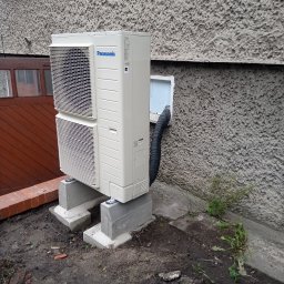 Instacjekompleksowo.pl- Tze.pl Wrocław - Zewnętrzna jednostka klimatyzatora Panasonic zamontowana na betonowych podstawach przy szorstkiej ścianie budynku, z widoczną czarną rurą odprowadzającą skropliny.