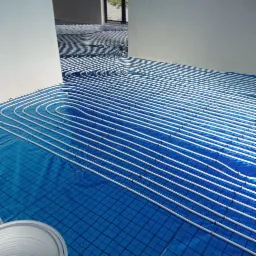 Białe rury ogrzewania podłogowego ułożone na niebieskiej folii z siatką, widoczne fragmenty ścian pomieszczenia.