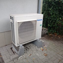 Instacjekompleksowo.pl- Tze.pl Wrocław - Zewnętrzna jednostka klimatyzacji Panasonic R32 ustawiona na betonowych bloczkach na kostce brukowej, z widocznym fragmentem ściany budynku i zieleni w tle.