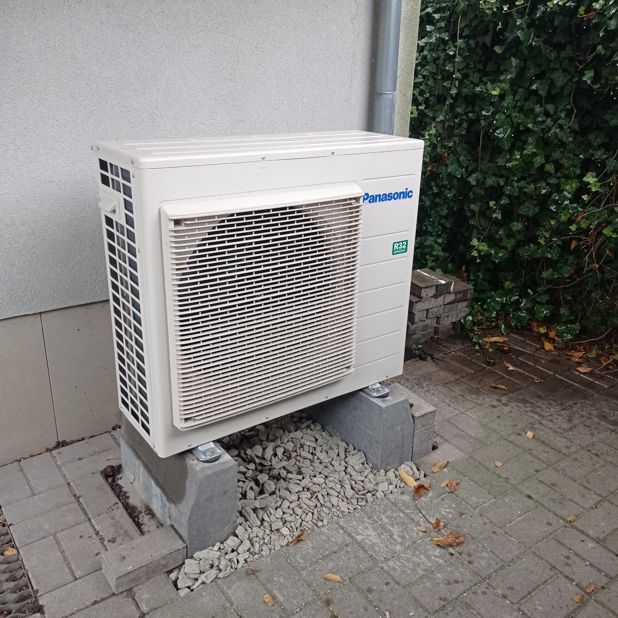 Zewnętrzna jednostka klimatyzacji Panasonic R32 ustawiona na betonowych bloczkach na kostce brukowej, z widocznym fragmentem ściany budynku i zieleni w tle.