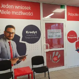 Wejście do biura oferującego wsparcie finansowe, z informacją o godzinach otwarcia i plakatami reklamowymi prezentującymi uśmiechniętych doradców i hasła o dostępności kredytów.