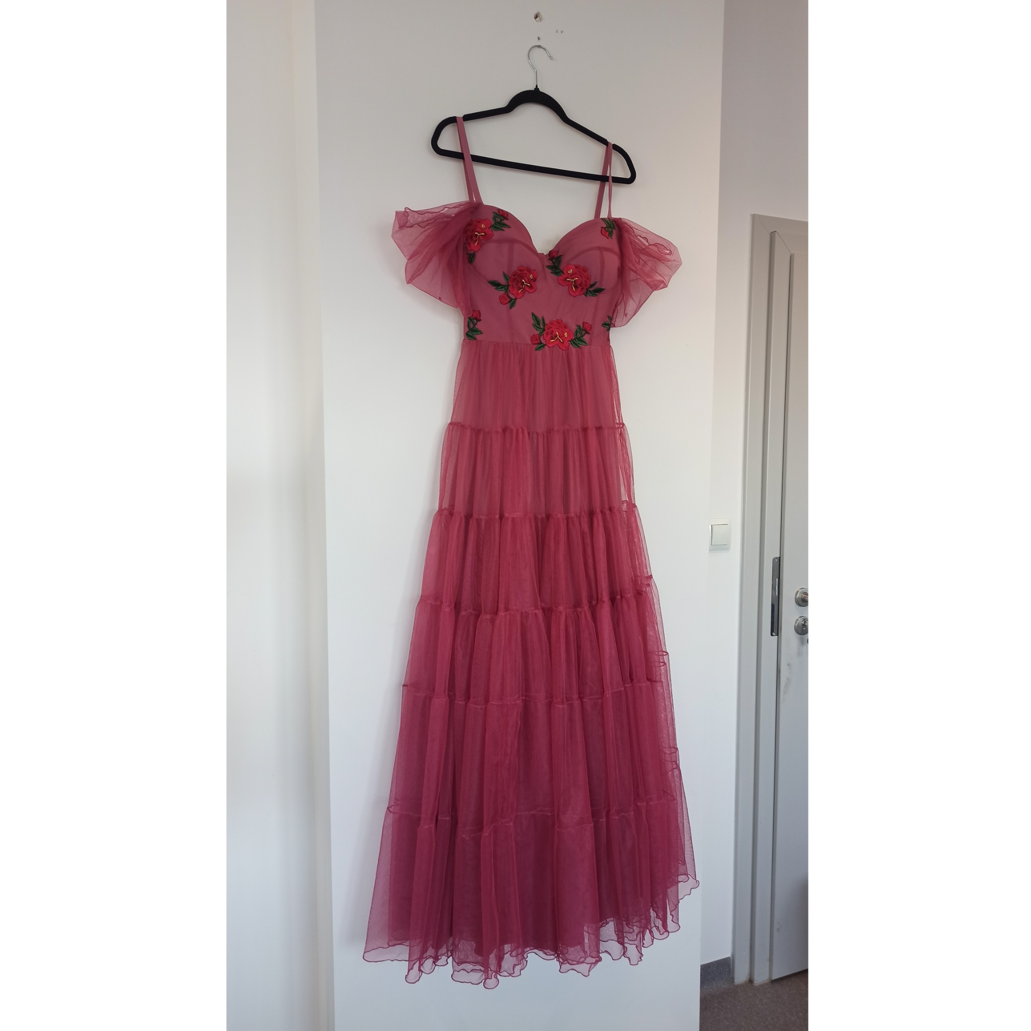 Długa, tiulowa suknia w odcieniu dusty rose, z warstwową spódnicą i aplikacjami z czerwonych róż przy dekolcie, wisząca na czarnym wieszaku na białej ścianie.