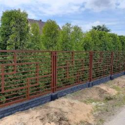Nowoczesna, brązowa konstrukcja ogrodzeniowa z geometrycznym wzorem, osadzona na podmurówce z szarej cegły, na tle zielonych tuj i częściowo widocznego dachu domu.