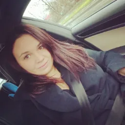 Kobieta z brązowymi włosami, uśmiechnięta, siedzi w samochodzie z zapiętym pasem bezpieczeństwa, widok przez okno na zewnątrz, w tle drzewa.