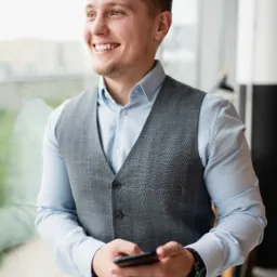 Uśmiechnięty mężczyzna w szarej kamizelce i błękitnej koszuli, trzymający telefon w dłoniach, patrzy w okno z rozmytym widokiem na miasto.