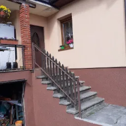 Dom z beżową elewacją, brązową podmurówką i schodami z drewnianą barierką. W oknie skrzynka z różowymi kwiatami. Widoczny fragment dachu z rynną i lampą.