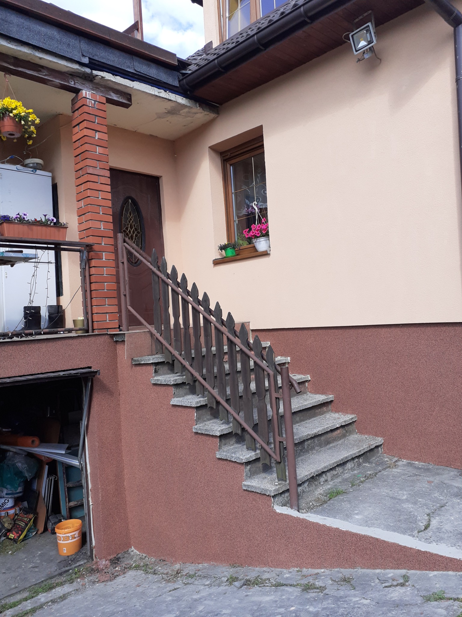 Dom z beżową elewacją, brązową podmurówką i schodami z drewnianą barierką. W oknie skrzynka z różowymi kwiatami. Widoczny fragment dachu z rynną i lampą.