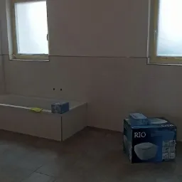 Wnętrze łazienki w trakcie prac, z widoczną zabudowaną wanną, częściowo wyłożoną jasnymi płytkami, oraz pudełkami po ceramice sanitarnej na podłodze z szarych płytek. Okna zabezpieczone folią...