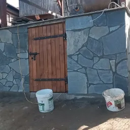 Zewnętrzna ściana budynku obłożona nieregularnym kamieniem w odcieniach szarości z drewnianymi drzwiami z metalowymi okuciami, widoczne dwa wiadra po farbie oraz fragment terenu przed budynkiem